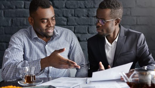 Attractive African businessman in glasses and suit holding papers, showing presentation of his project to cheerful business partner, who smiling happily, supporting his idea with positive gesture